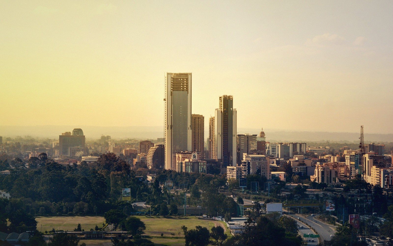 Tall skyscrapers rise above Nairobi's skyline at sunset, with green parkland in the foreground
