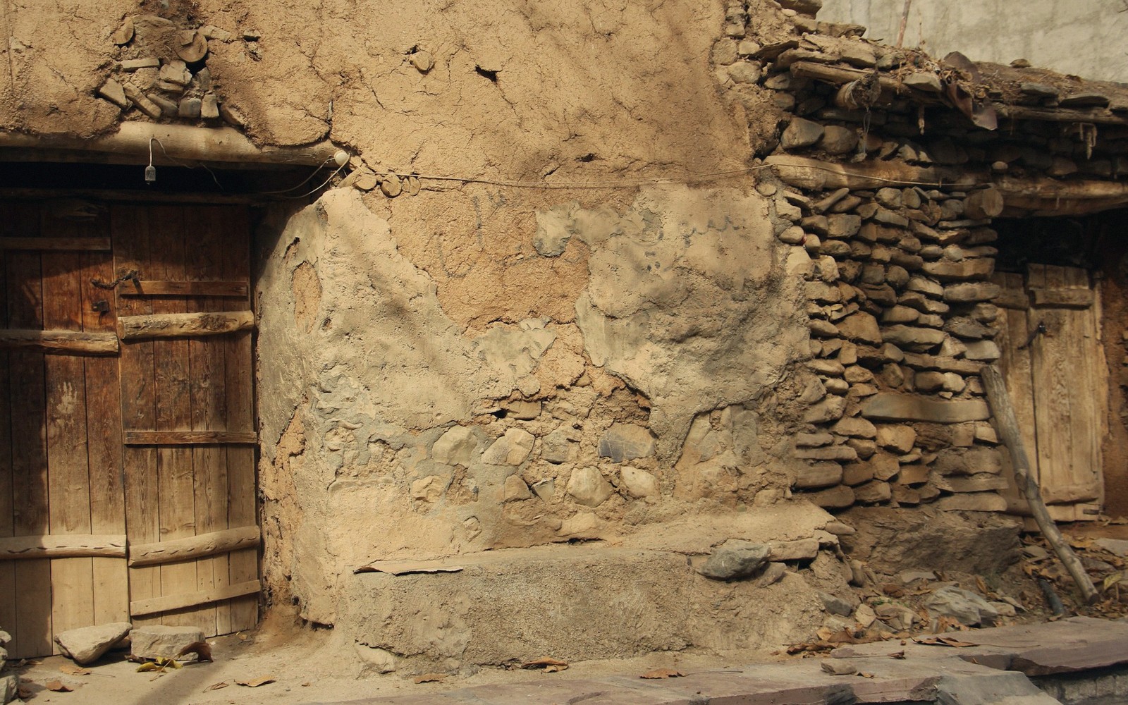 Weathered mud-plastered wall with wooden doors and exposed stone masonry